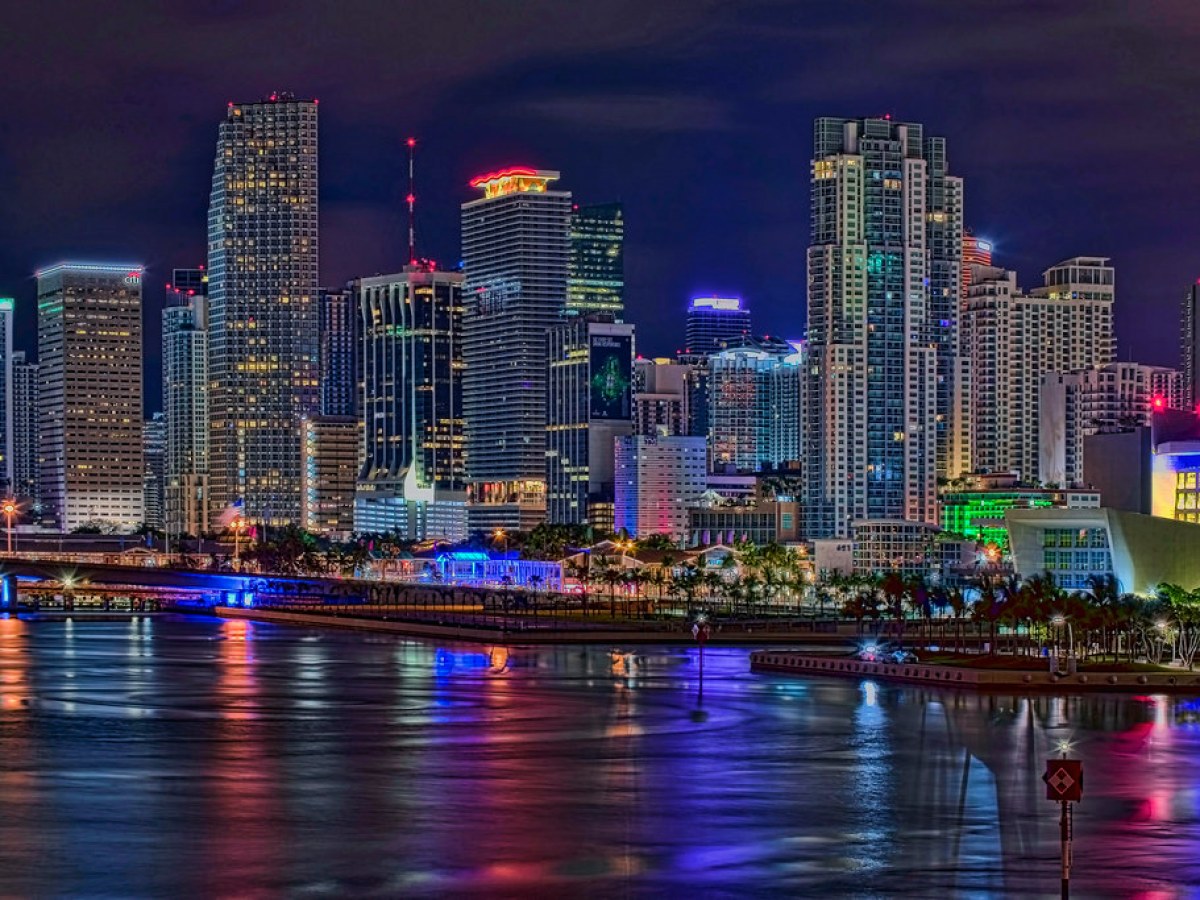 Colorful city skyline at night reflected in water, with tall lit-up buildings.